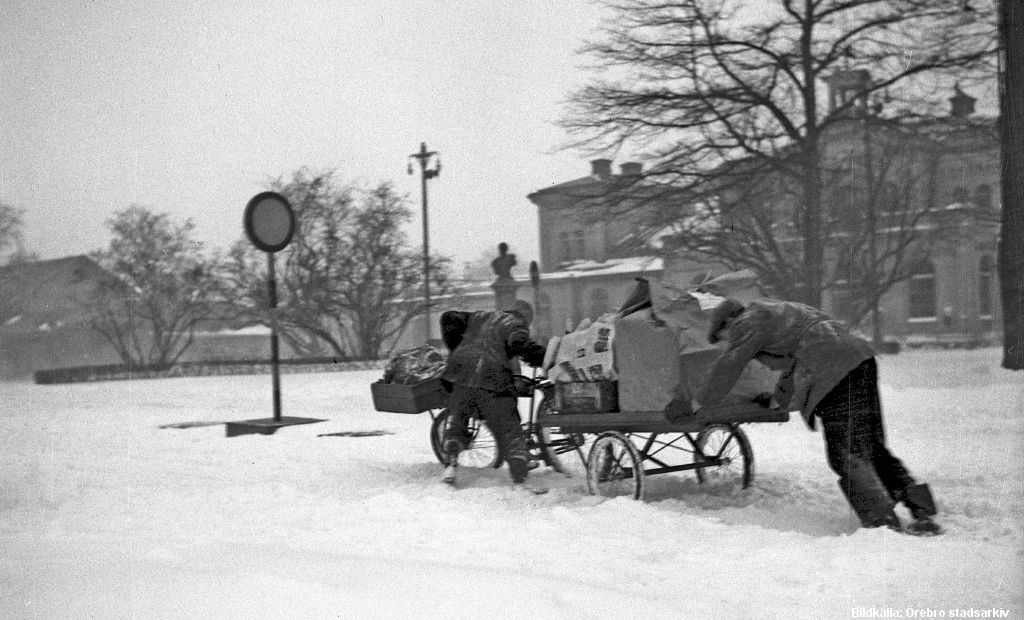 Två män knuffar en packcykel med mycket last i snöoväder. Tung transport till Örebro Centralstation, december 1945. Foto Specialfoto AB/Örebro stadsarkiv.