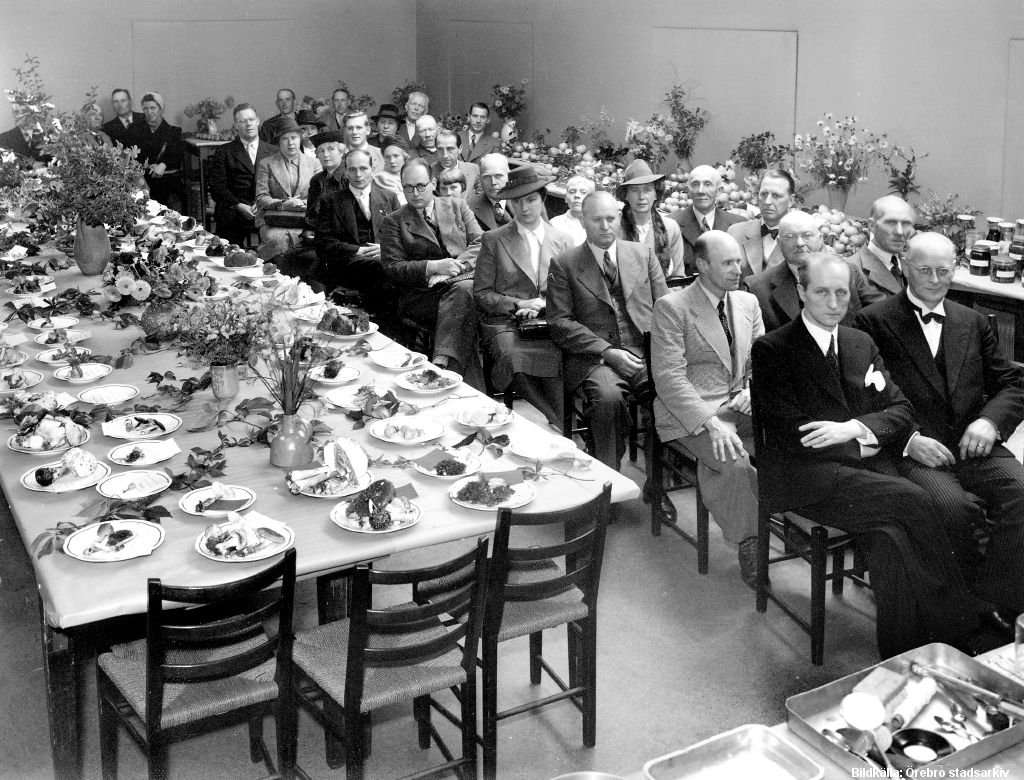 Ett stort matsalsbord med tallrikar med svamp. Vid bordets långsida sitter människor i tre rader. 