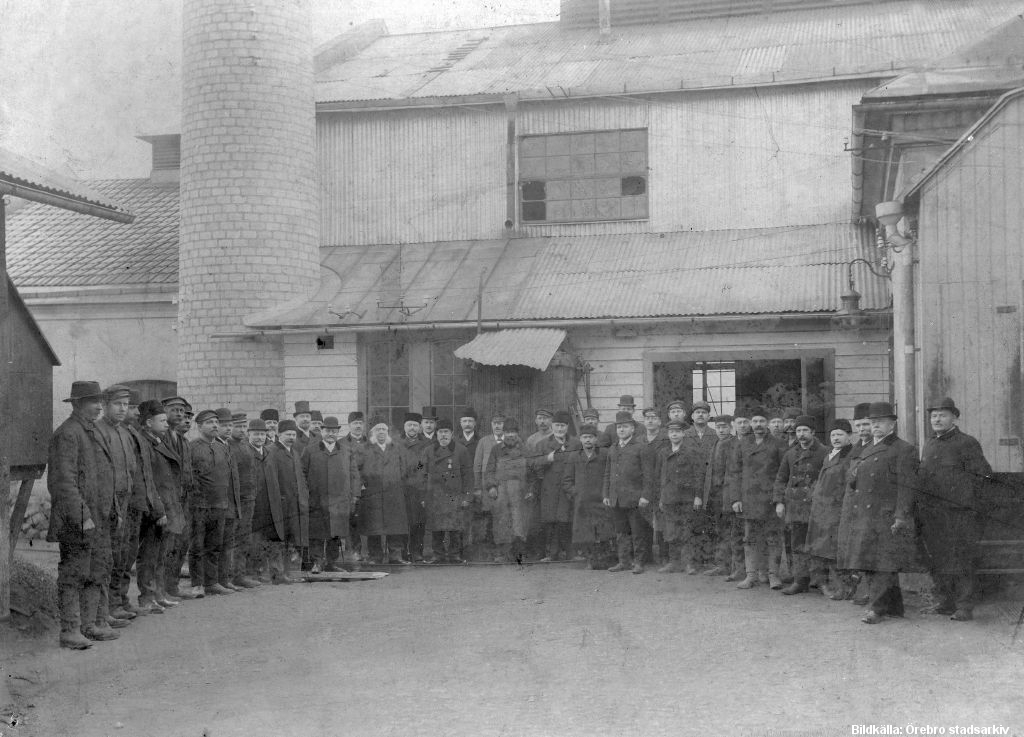 Svartvitt fotografi på ett 40-tal män i hatt och långa rockar. De står uppradade framför en industribyggnad.  