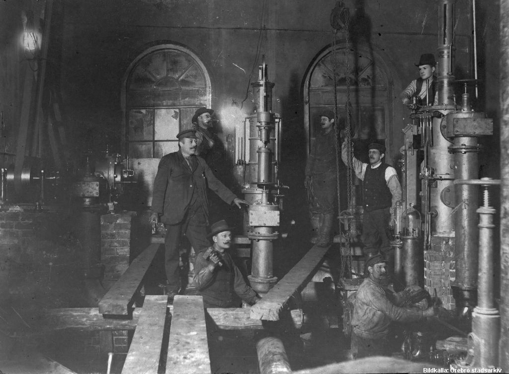 Sex män står vid maskiner i ett mörkt rum.