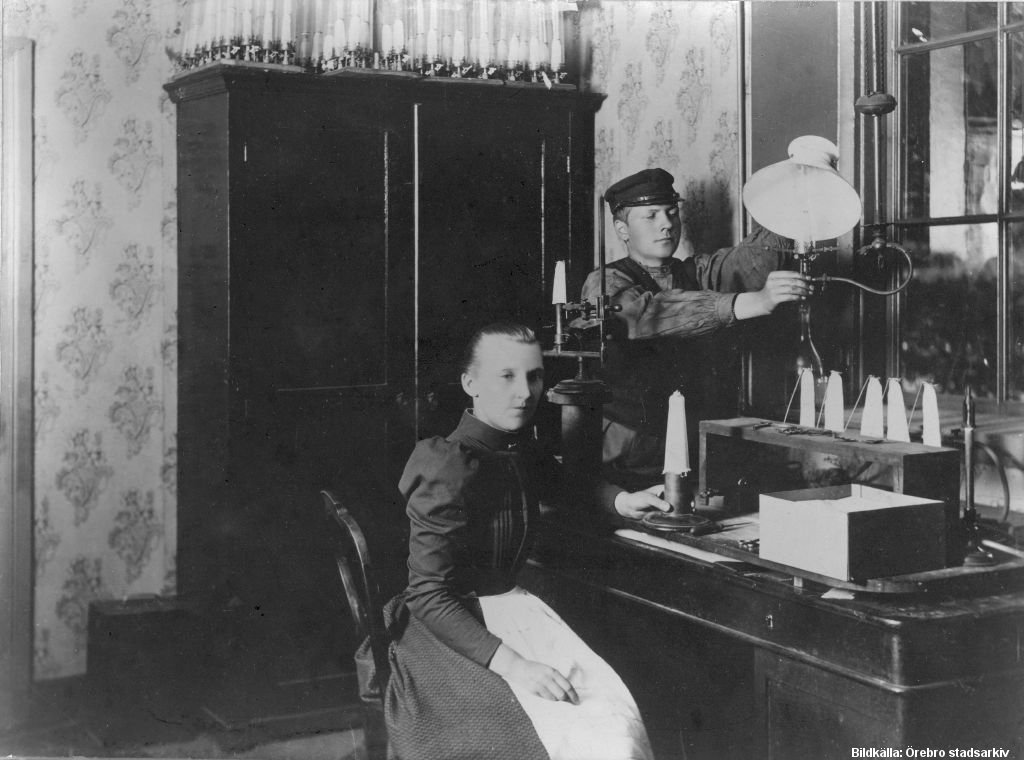 Kvinna sitter vid bord med lampor. En ung man står brevid, hållandes en lampa.