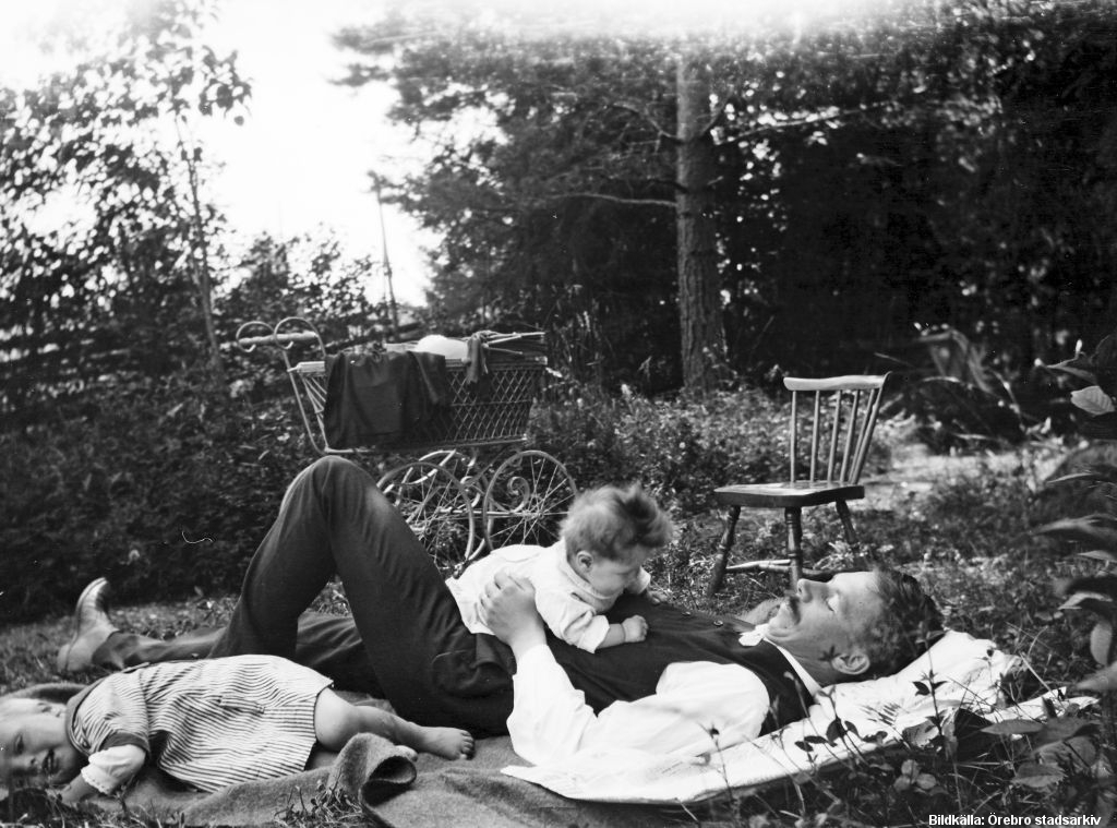 Man och två barn vilar på en filt i en trädgård.