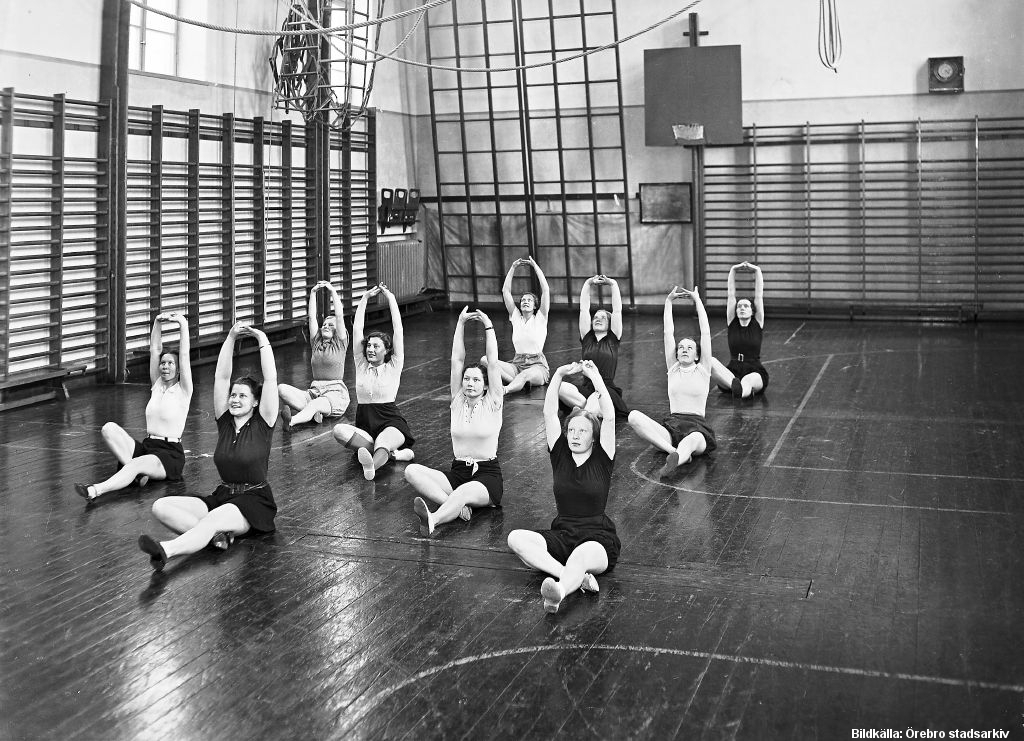 I en gymnastiksal med ribbstolar längs väggarna sitter en grupp unga kvinnor och gör samma rörelse, med benen i kors och armarna över huvudet.