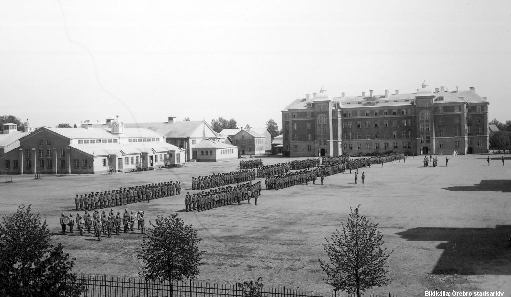 Militärer står uppradade på gård framför stort fyravåningshus.