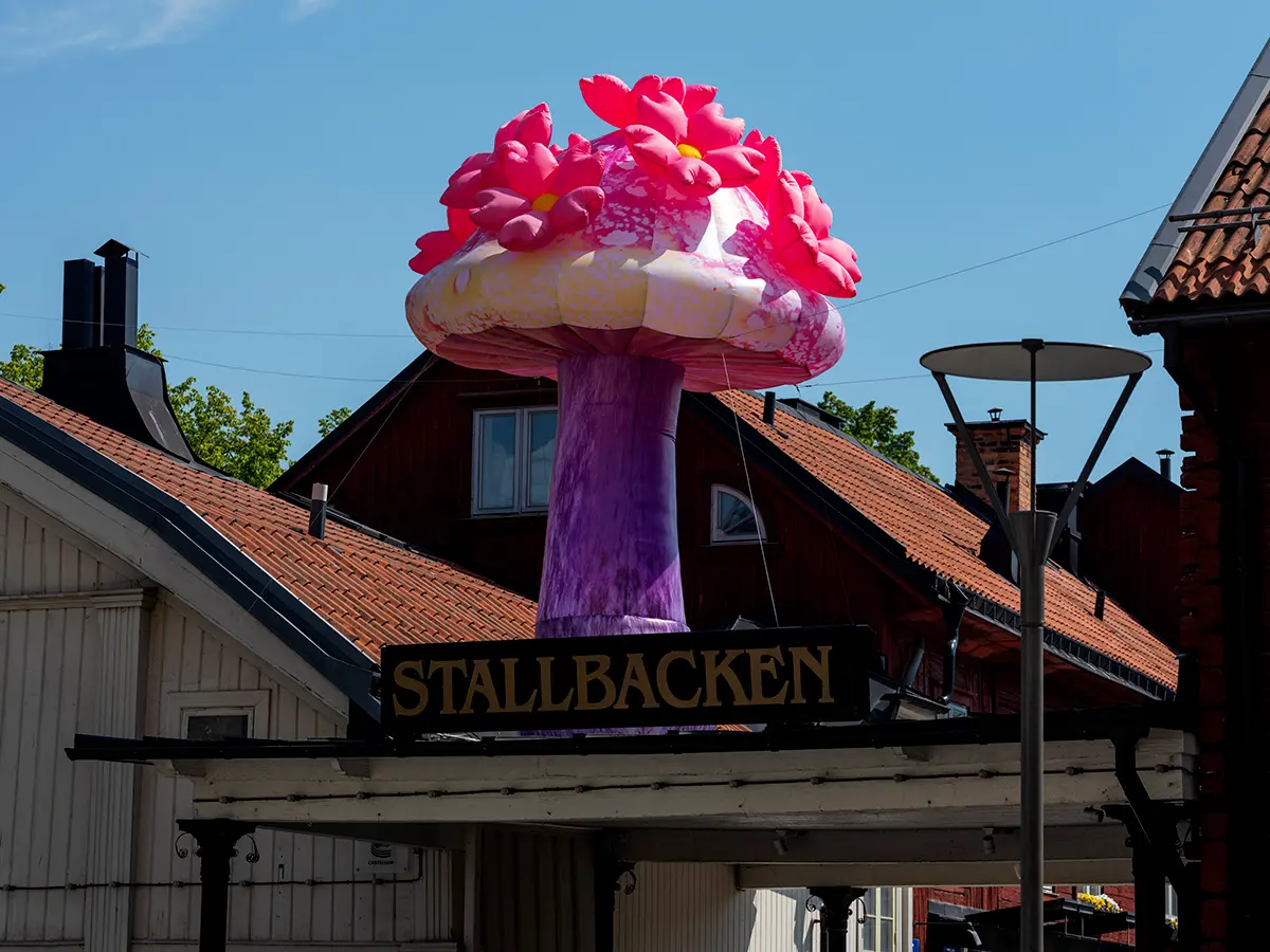 En uppblåsbar svamp i rosa och lila färger med blommor på toppen, syns ovanpå hustak i gammal stil. På en skylt på taket står det Stallbacken. Himlen bakom är blå.