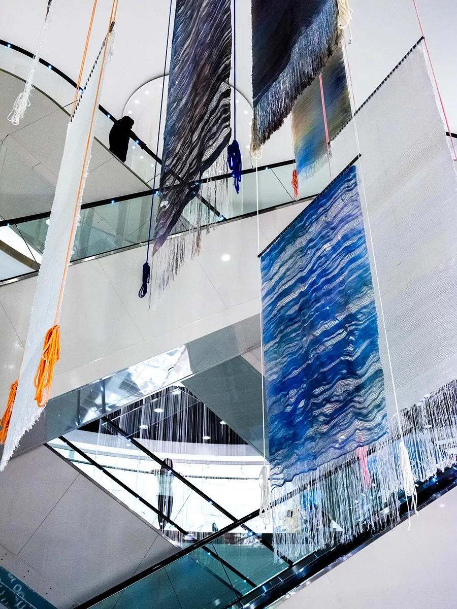 I utrymmet mellan rulltrapporna i en galleria hänger vävar ner från taket i ljusa färger