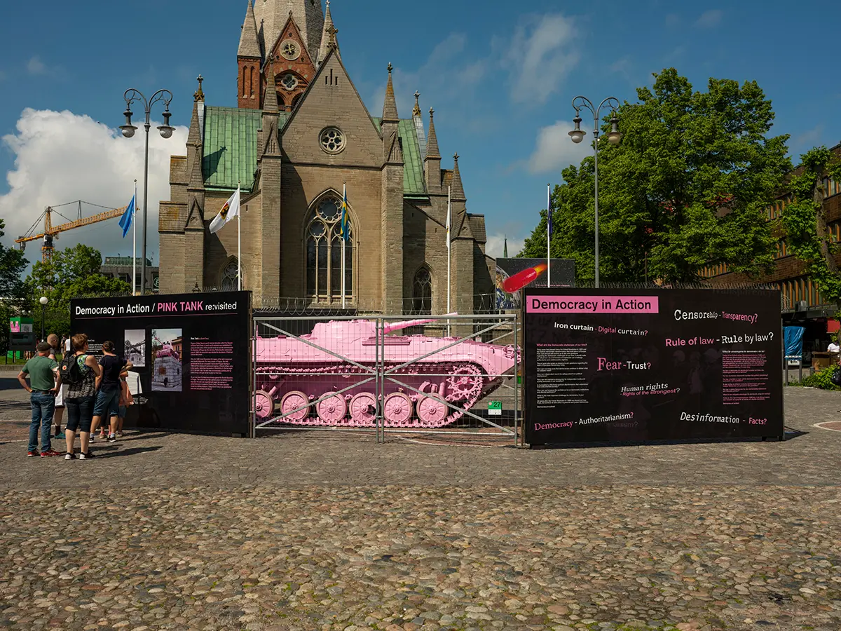I en inhängad framför en kyrkbyggnad står en rosa stridsvagn, på sidorna av inhägnaden sitter svarta paneler med rosa och vit text. Det står en grupp människor och läser på panelerna.  