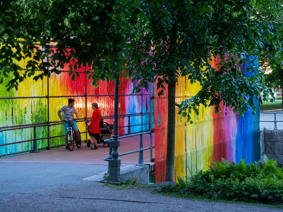 Två personer på cykel har stannat på en bro som blivit klädd i remsor gjorda av regnbågens alla färger av färger