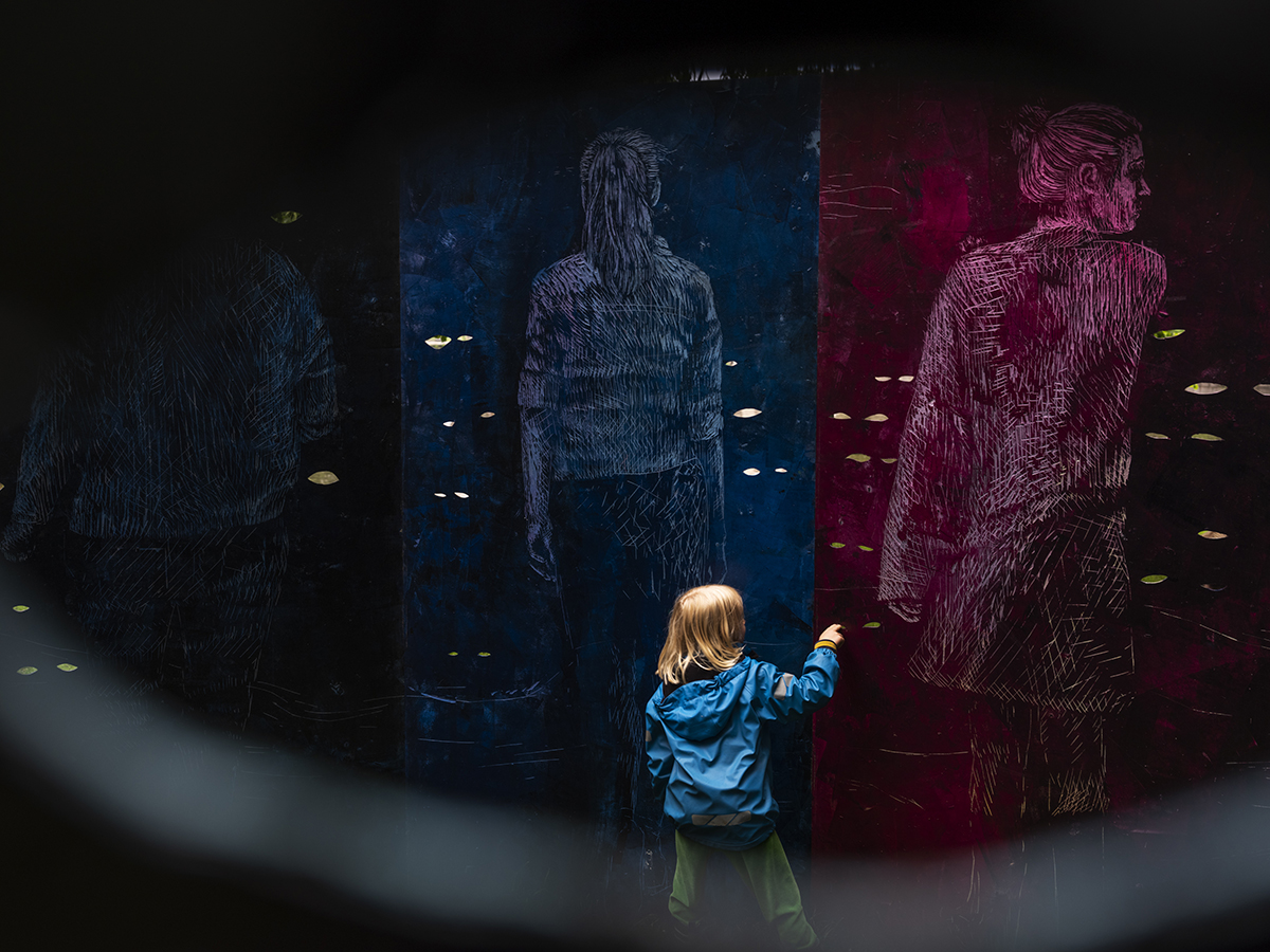 Ett barn med ljust hår och blå jacka har lyft sin hand mot två stora teckningar på en vägg. Den ena teckningen är rosa/lila och föreställer en kvinna bakifrån i helkroppsfigur som vänder bort huvudet mot höger.  Den andra teckningen är blåaktig och föreställer en annan kvinna bakifrån i helkroppsfigur.