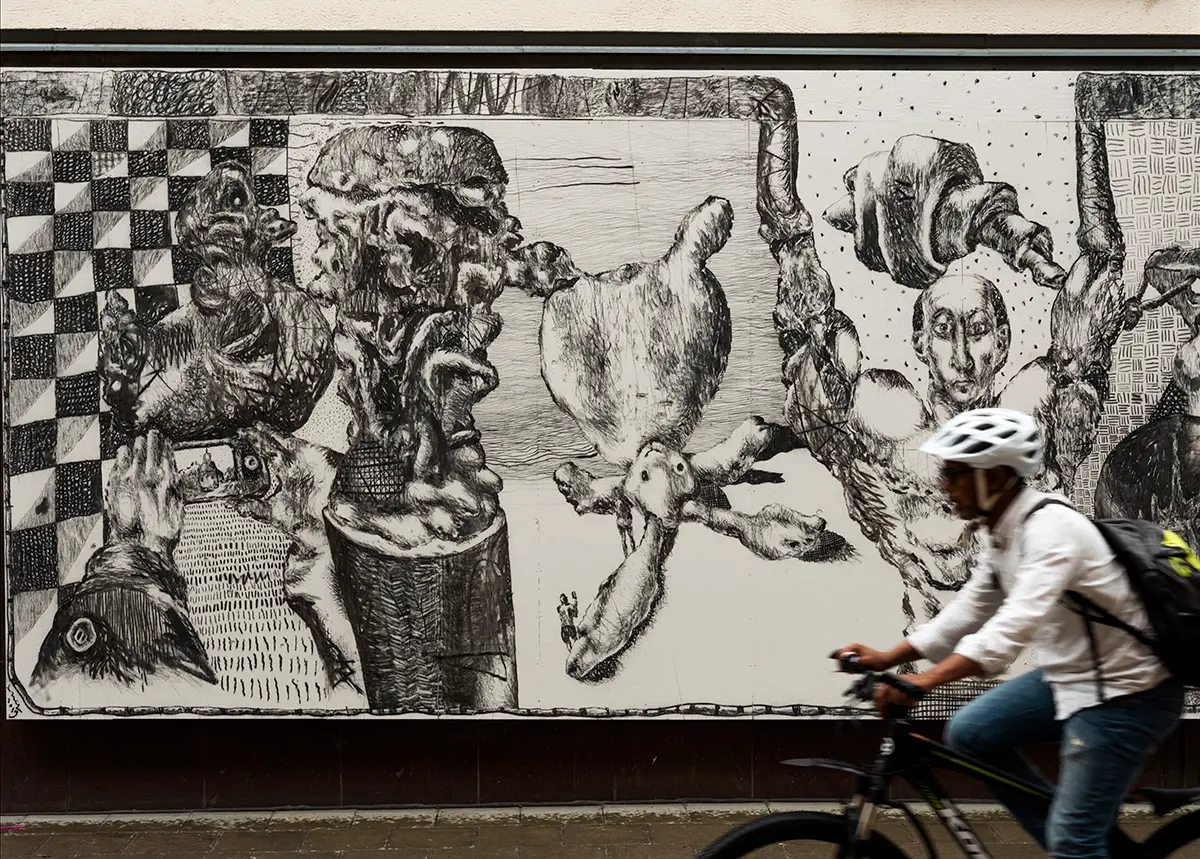 En avlång väggmålning sitter uppsatt längst ner på ett vitt hus. Bilden är svartvit och i teckningsstil och föreställer flera olika motiv, däribland en kanin, en anka, en mobiltelefon och en människa som sträcker upp armarna. I bakgrunden finns olika abstrakta mönster. En man cyklar förbi på gatan framför bilden. 