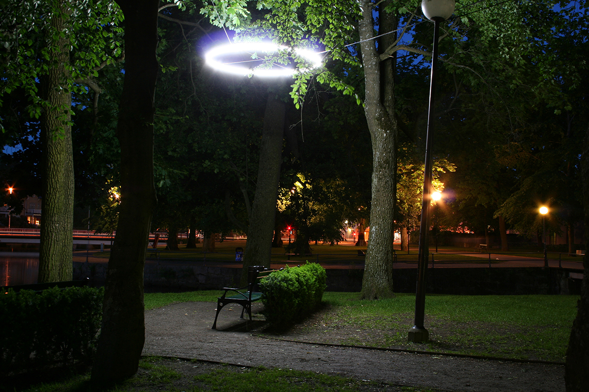 Det har blivit kväll och det är mörkt ute men i slottsparken lyses en parkbänk upp, över den sitter ett runt lysrör som en gloria som lyser starkt nedåt. 