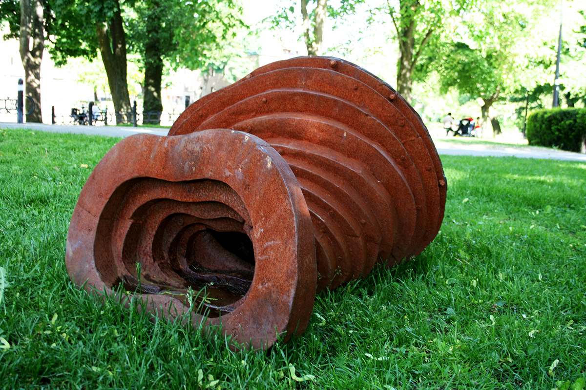 Utomhus på en gräsmatta ligger en rostig skulptur av sammansatta vågformade ramar i olika storlekar. 