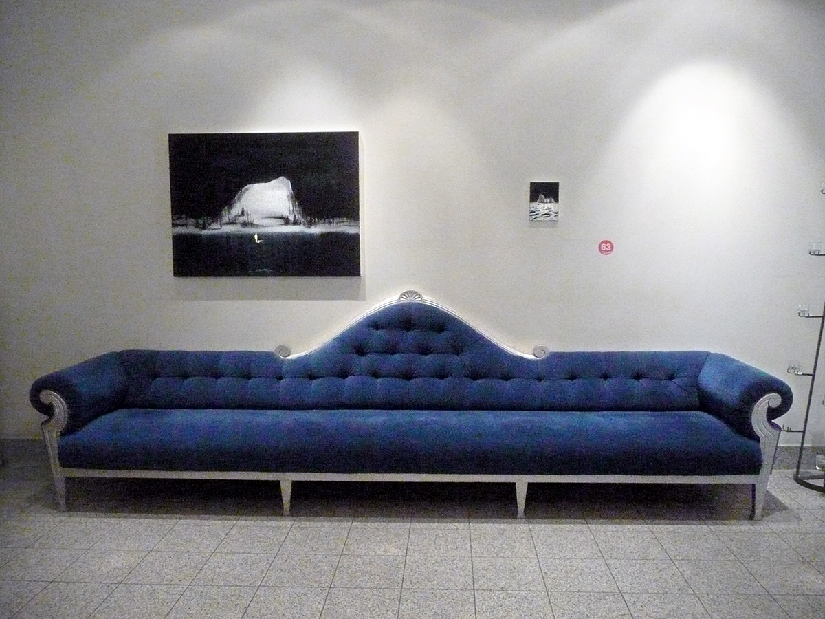 Mot en vit vägg står en lång blå elegant soffa. På väggen hänger svartvita konstverk av landskap med snö och is. 