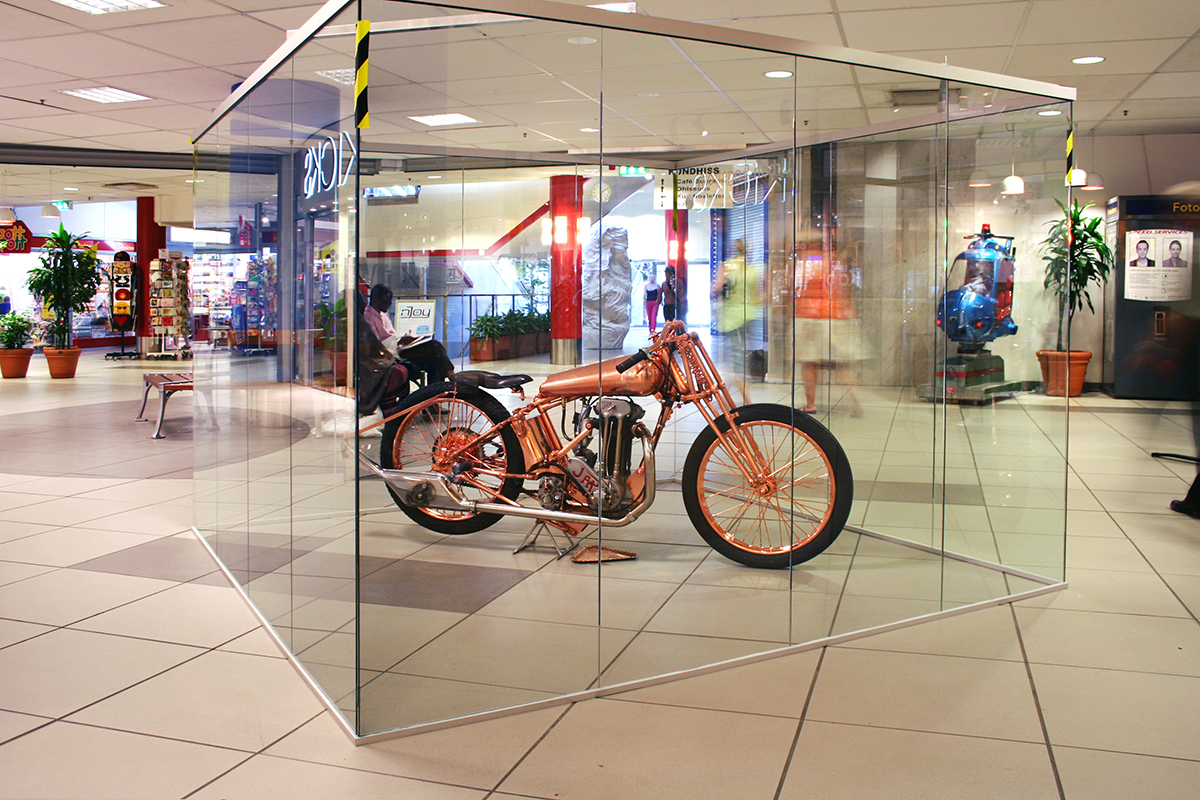 Inuti en glasmonter står en koppar färgad motorcykel. 