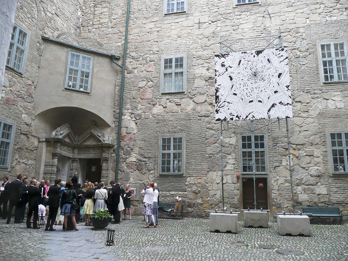Vid Slottets ingår står två betongsuggor och två byggställningar hänger en svartvitt målning uppe i luften. Målningen är målad i ett spindelnät mönster. 