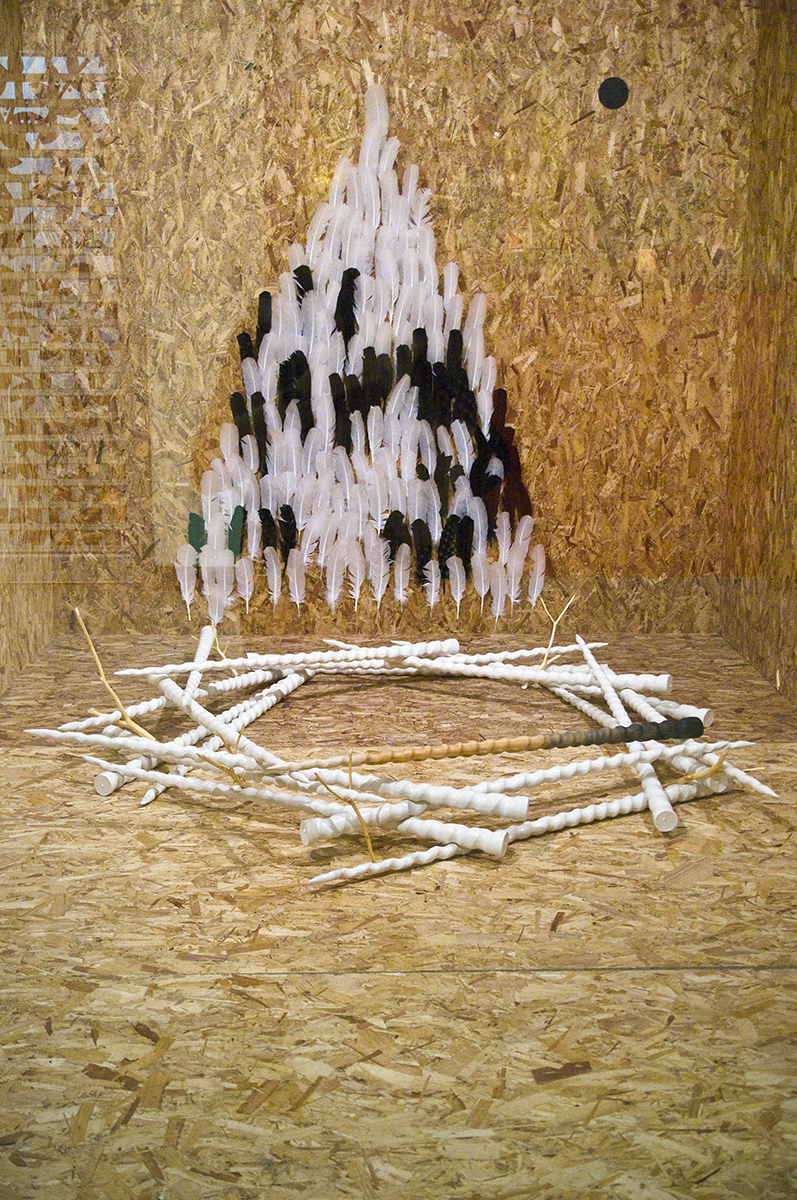 Inuti en glasmonter med plywood väggar sitter svarta och vita fjädrar på väggen som en triangel och på golvet ligger vita enhörningshorn på varandra som formar en sexhörning.