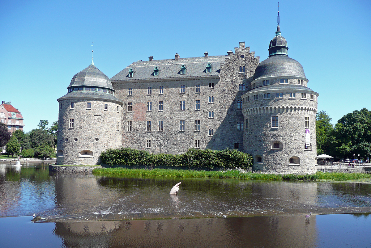 Framför slottet sticker en verklighetstrogen överdimensionerad hand upp ur vattnet som pekar med ena fingret mot höger.