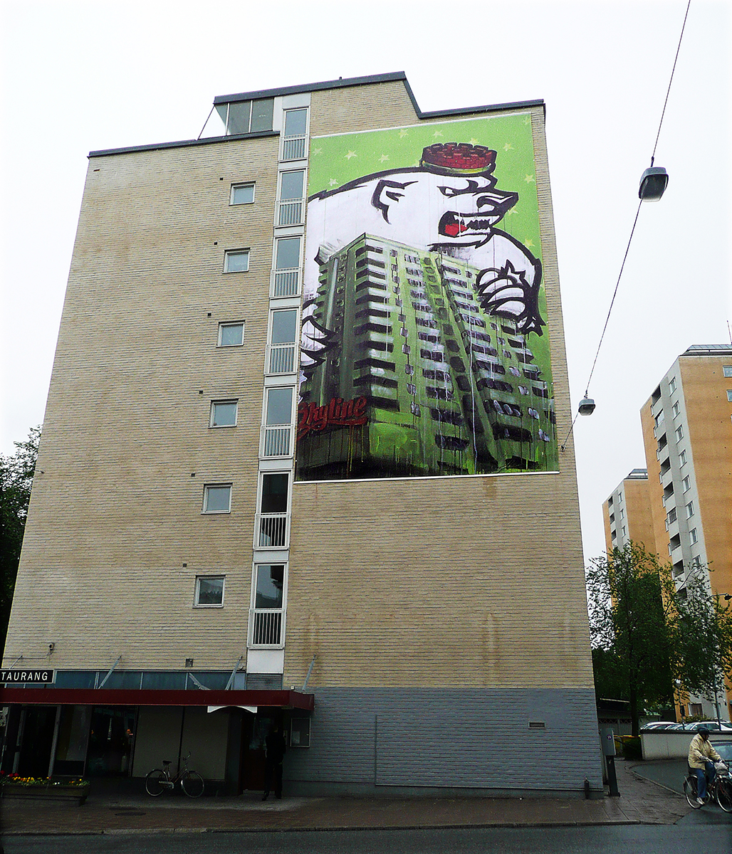 På en husfasad hänger en vepa av en målning som innehåller ett fotografi av ett högt lägenhetshus som en isbjörn attackerar. Isbjörnen har en röd hatt. 