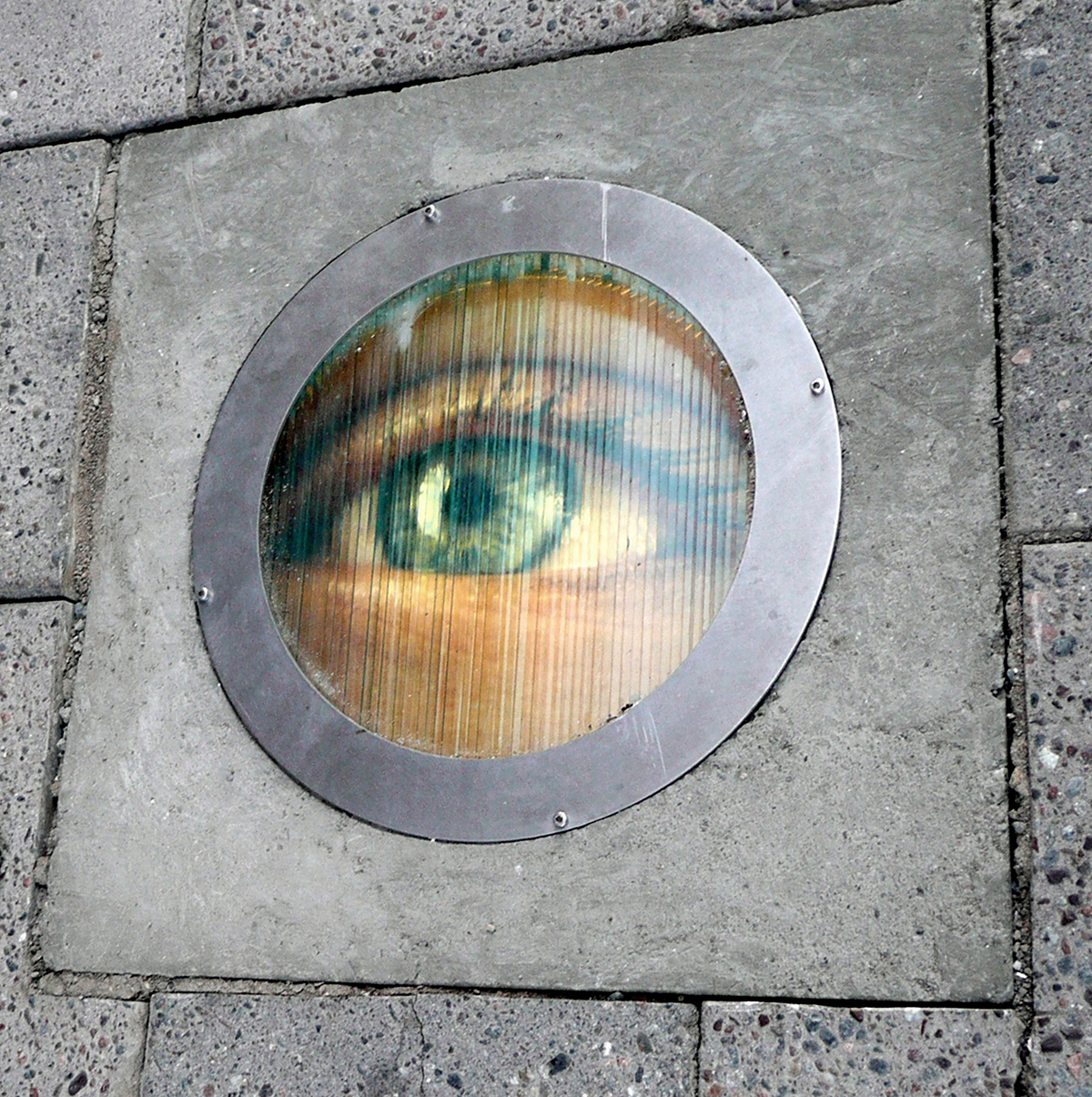 Ett öga är målat på ett runt gasskydd för en gatlampa som sitter i marken.  