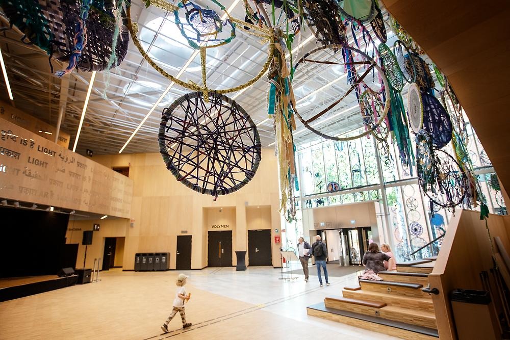 In the foreground hang rings covered with textiles in blue colors. In the background you can see a room and people walking by.
