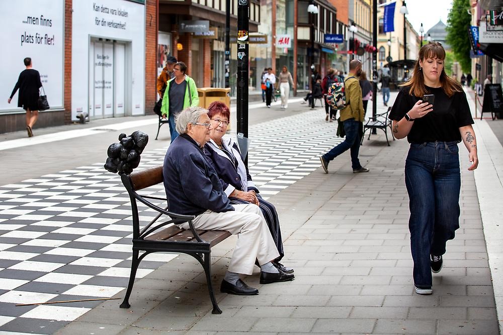 A man and a woman are sitting on a park bench in an urban environment. A sculpture is placed on the backrest. A woman walks past in front of them. The sculpture is approximately 30-40 cm high and is made of tightly wrapped black textile.