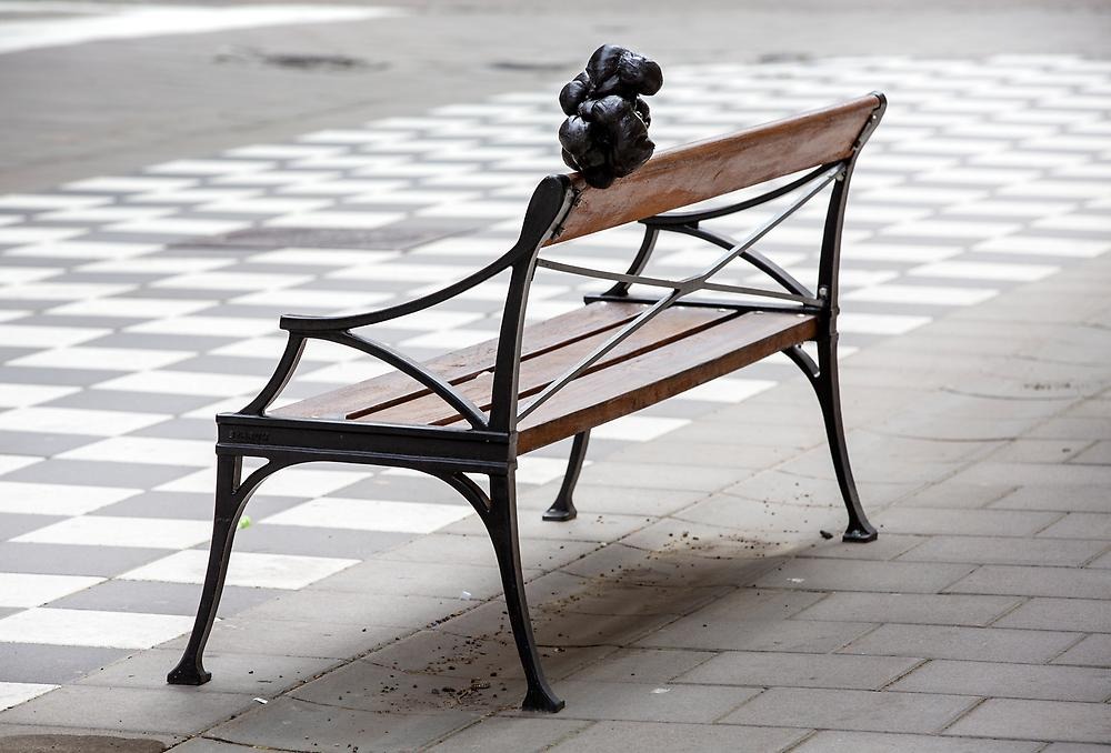 On an empty park bench on a street, a black object balances on the backrest. The object is a black "lump" of wrapped fabric.