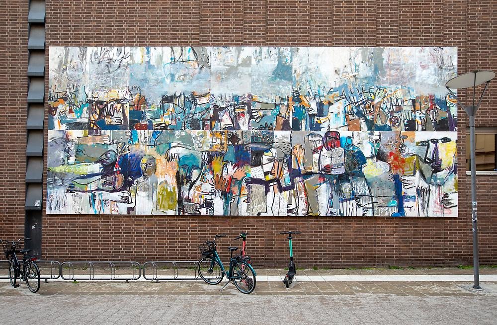 A large abstract painting is placed on a brick wall. The colors are muted and blurry. One can sense faces and bodies in the multitude of color and shape.