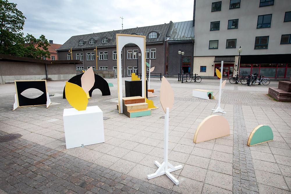 In a square stands a playful installation of modules made of wood. Some are pastel and some are black. The modules have the shape of semicircles, columns with leaf-shaped parts and a doorway.