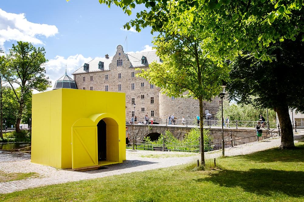 A yellow-painted small house with an arched door is placed on a ledge that looks out over the water, in the background you can see Örebro Castle, in the foreground you can see grass and trees.