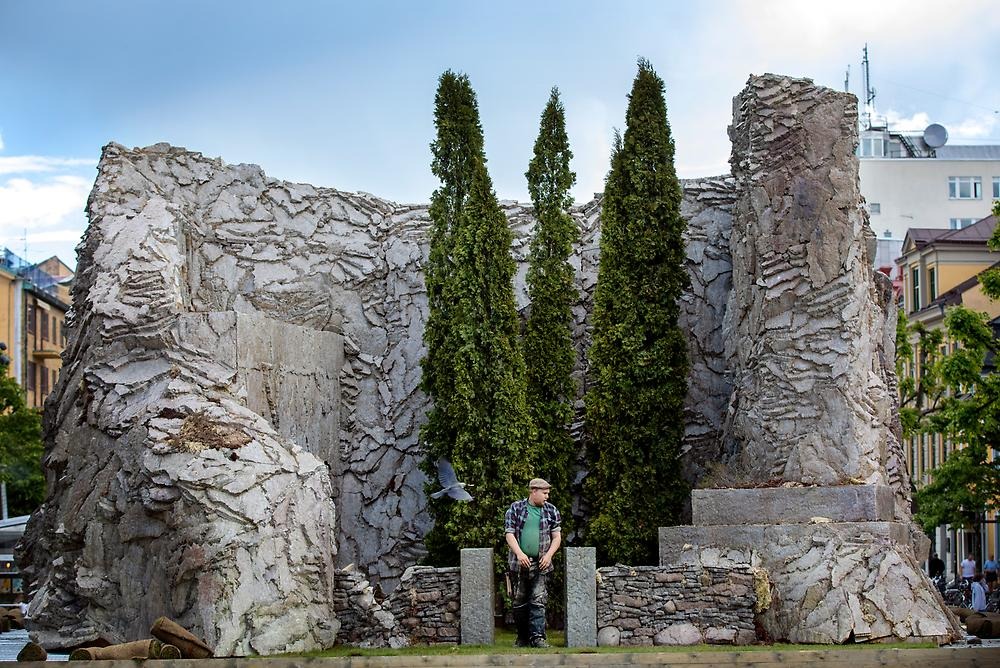 Outside a monumental work of art stands in a square. It is shaped like a U with rock walls. In the center of the structure stand three trees (thuja). In the opening of the U, two pillars are placed, they also appear to be made of stone. Between the pillars you can see a man who is in motion, it is the artist Henrik Jonsson.