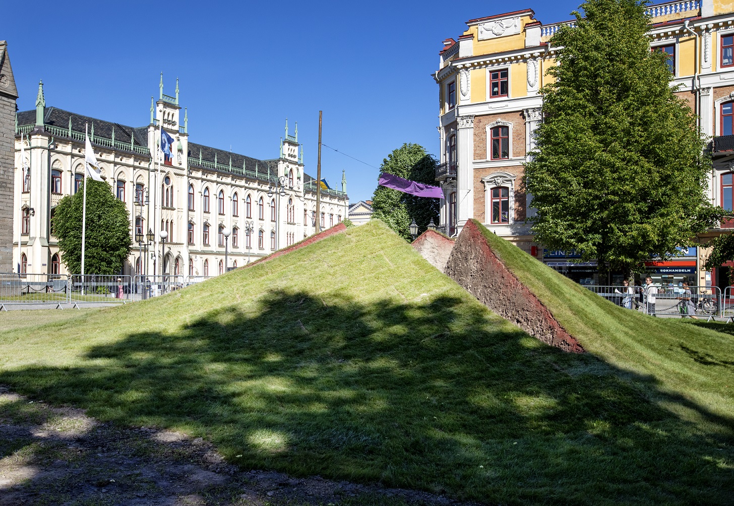 The lawn in front of Saint Nicolai Church has opened up like a wound from the underground. The work is made up of earth and grass that break up from the ground in small pyramid shapes that create a hole in the middle. In the background you can see the town hall.