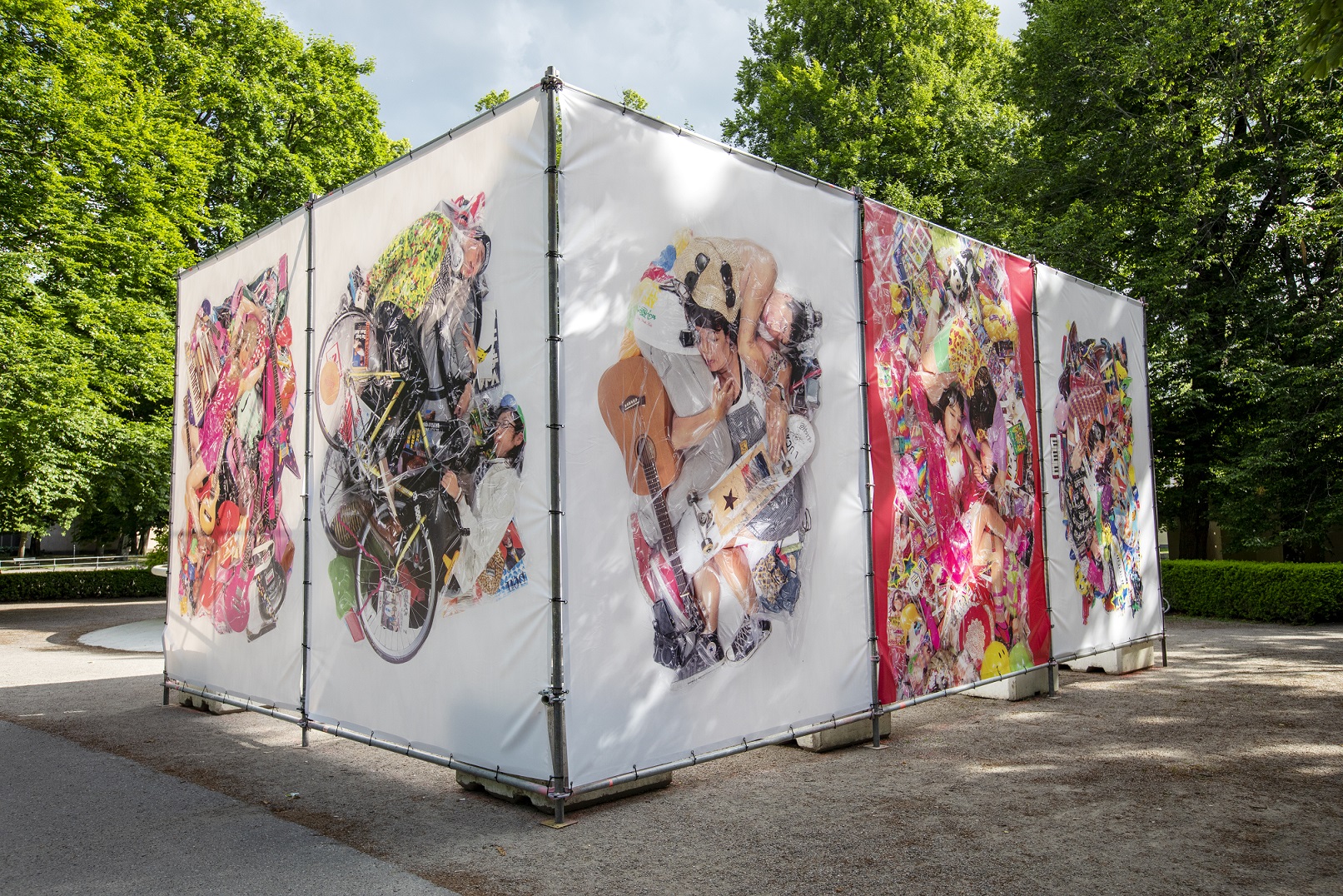 In a park there is a large stand that holds up five large photographs. Four pieces with white background and one with dark pink. The photos consist of people and everyday items that have been vacuum packed together.