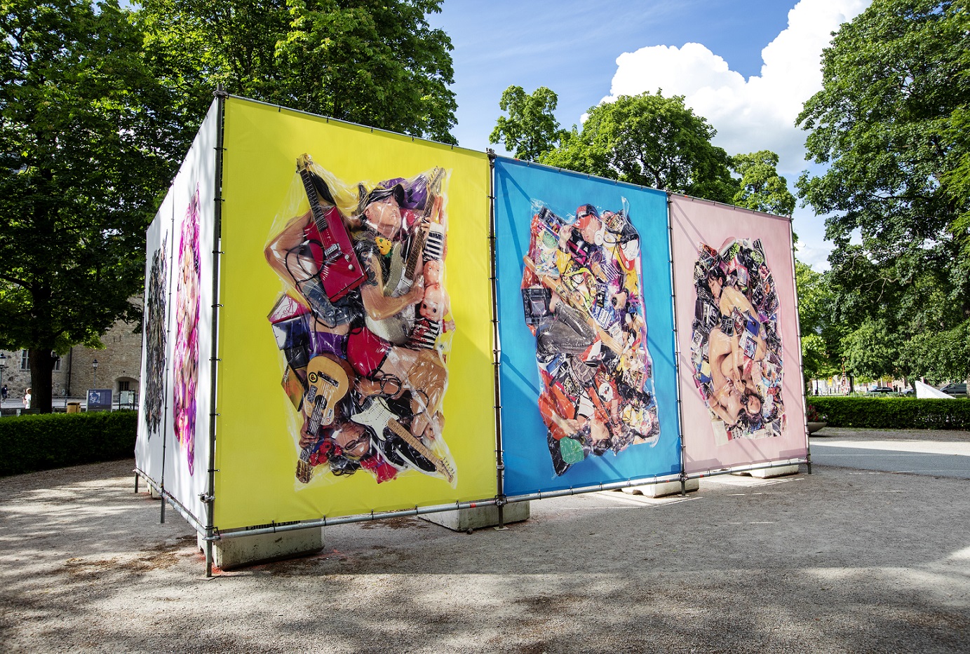 In a park there is a large stand that holds up five large photographs. Two images have a white background and the other three have different strong background colors yellow, blue and pink. The photos consist of people and everyday items that have been vacuum packed together.