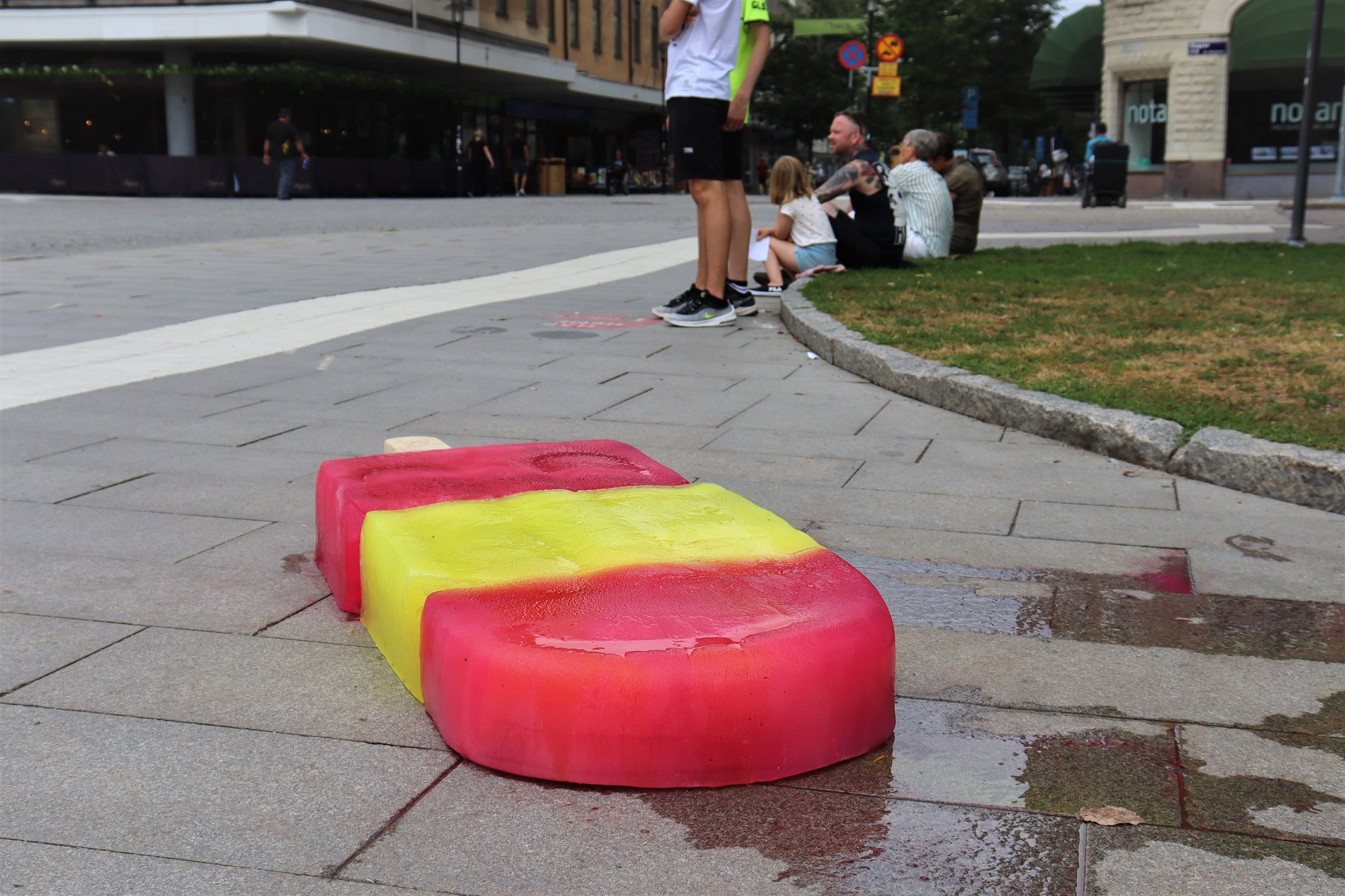 A yellow-red striped oversized two-meter-long popsicle with popsicle stick lies outdoors on the stone slabs, melting. This work of art was greatly appreciated by all the thirsty dogs that walked by.