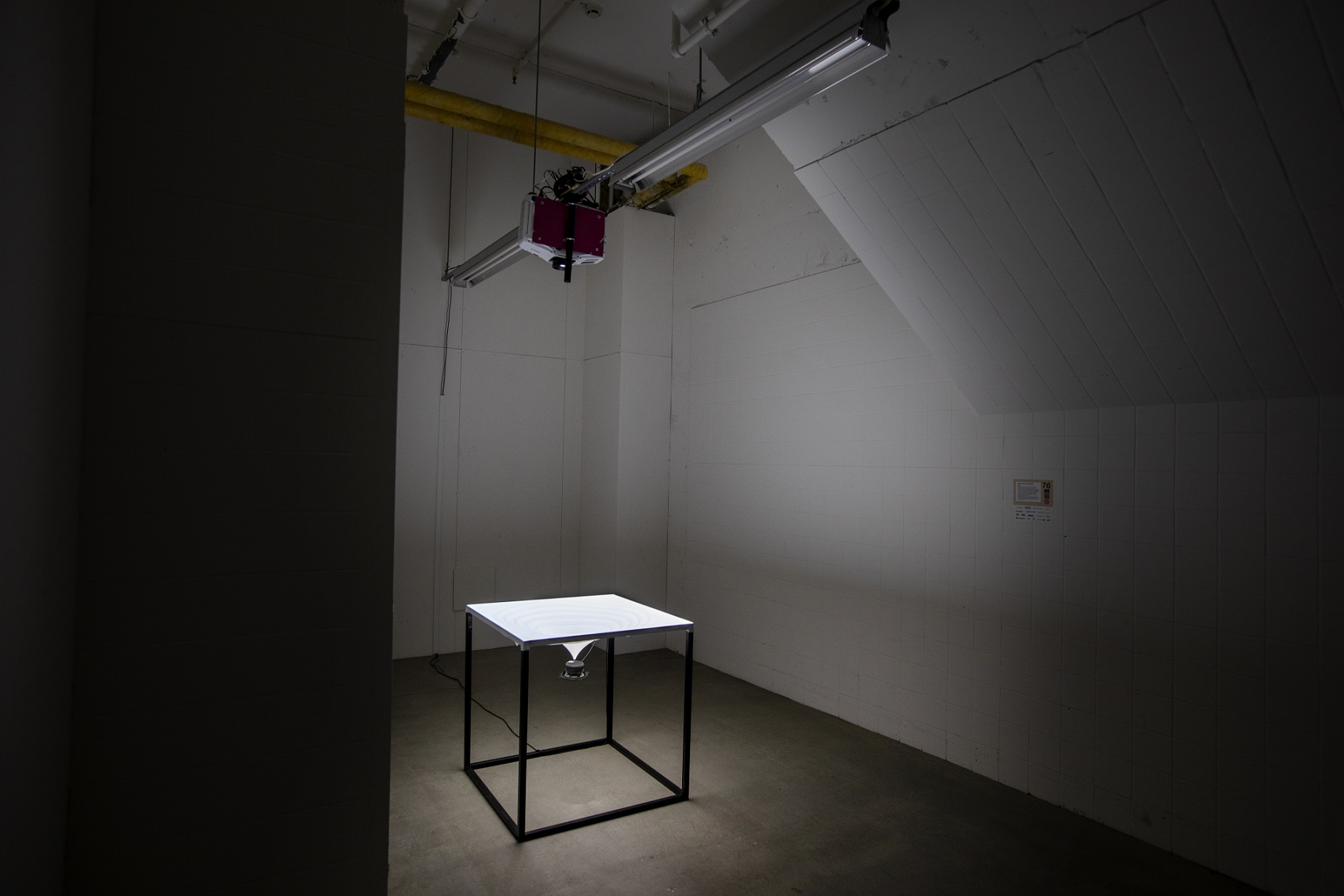 In the corner of a dark room stands a white table with a depth in it, which means that the surface of the table becomes conformal down to and at the bottom sits a lens. A projector hangs from the ceiling above and shines down on the table.