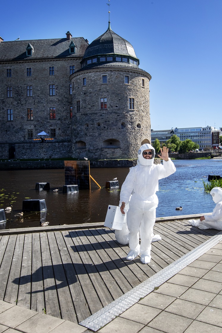 Two people dressed as astronauts in front of the castle. One sits on the wooden deck and one stands up and waves cheerfully at the camera.