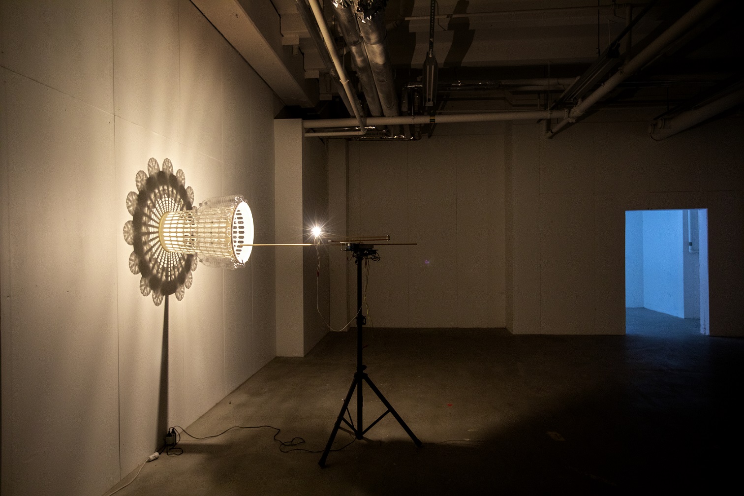 In a dark room stands a black stand with a light on it pointing at an object that looks like a tied plastic basket with plastic bottles attached to the outside. The light reflections turn into an mandala pattern that moves on the wall.