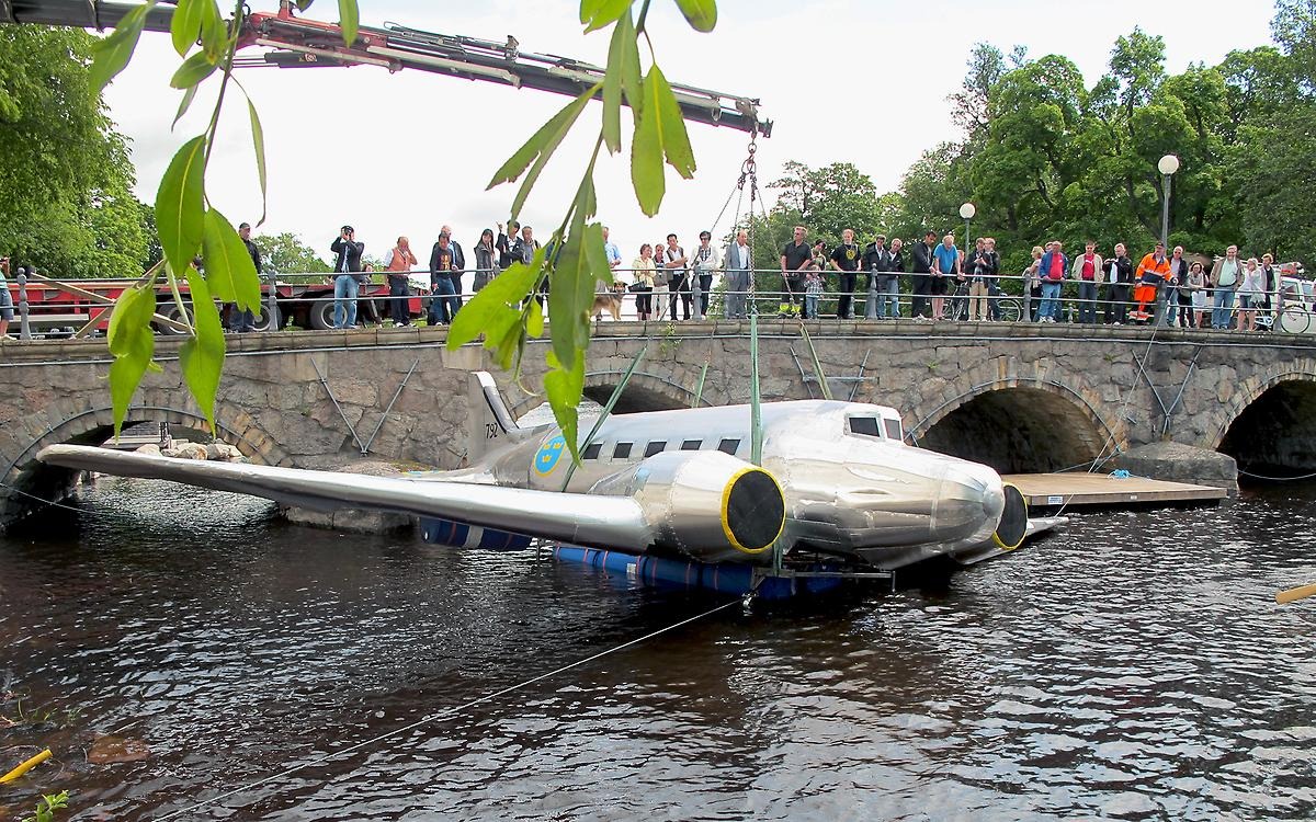 En bro framför Örebro Slott är fylld med människor som tar bilder mot vattnet där ett passagerarplan placeras ner i vattnet med hjälp av en kranbil. 
