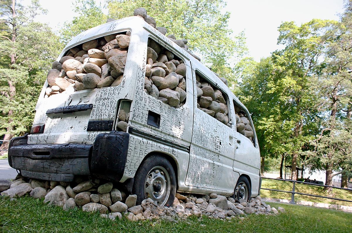 En vit minibuss utan glasfönster står på en hög med stenar samt är även helt fylld med stenar.