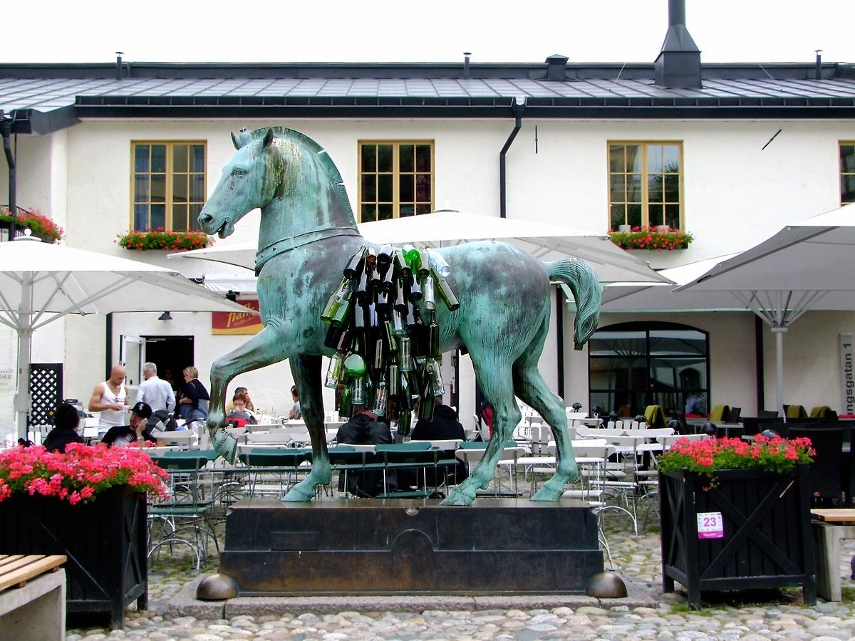 På ett torg framför en uteservering står en grön hästskulptur och på sidan av hästen sitter många glasflaskor i gröna färger fästa på varandra. 