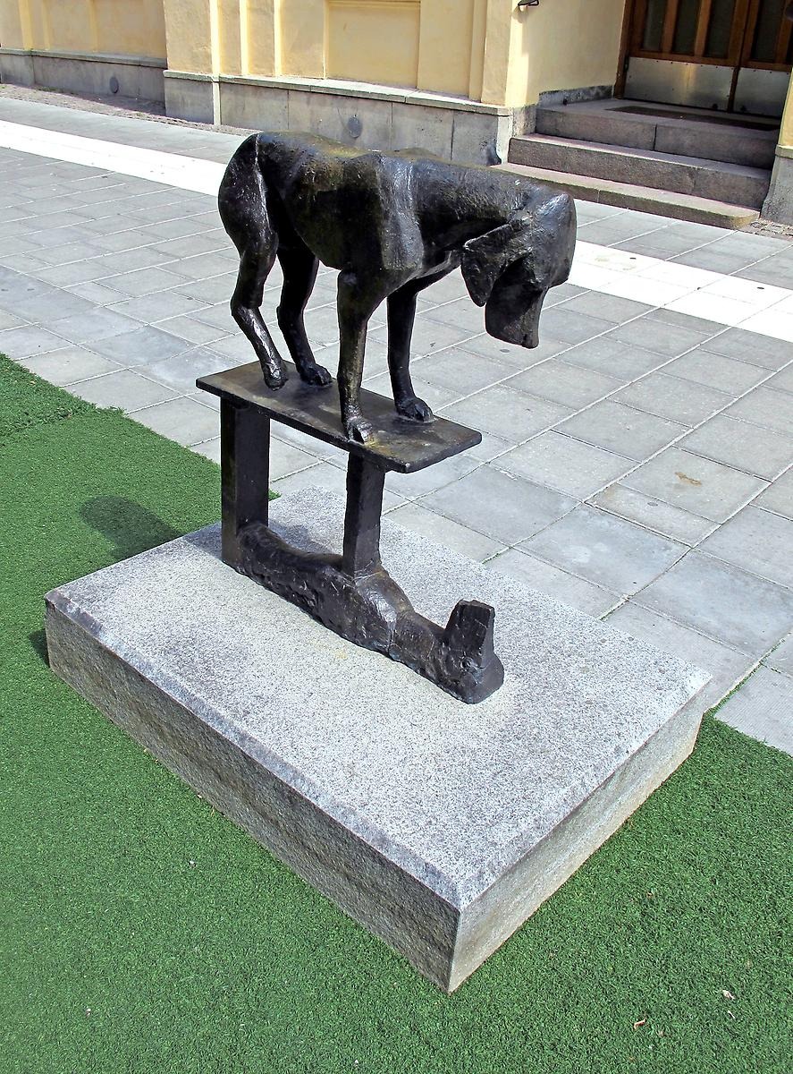 På en betongskiva står en svartfärgad staty av en hund som kollar ner och ser sin egna reflektion.