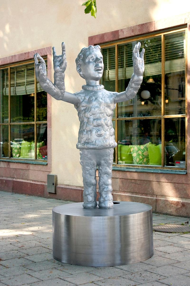Utomhus på stenplattorna står en aluminiumfärgad cylinderform samt på den står en silverfärgad staty av en pojke med tre armar. 