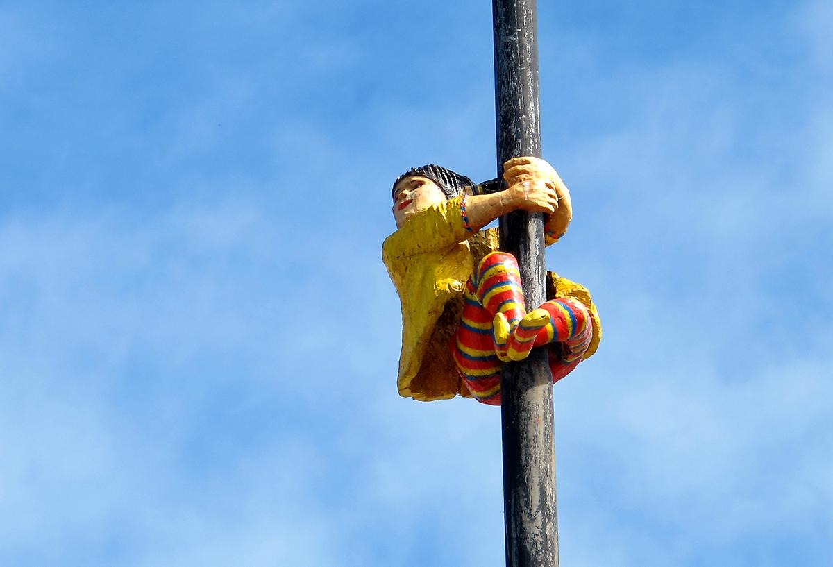 På en svart lyktstolpe har ett ett barn klämt sig fast, barnet är klätt i en gul jacka och randiga strumpbyxor i färgglada färger. 