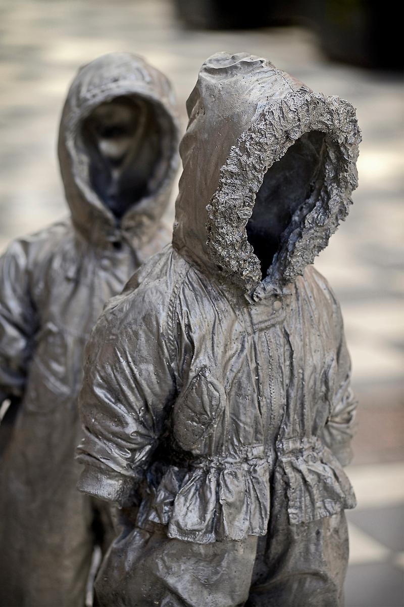 Skulptur av två gråbrunaktiga vinterjackor som står på rad. De har formen som att de är fyllda och att någon har på sig jackorna men där man vanligtvis ser ett människohuvud sticka upp är det bara ett mörkt hål.