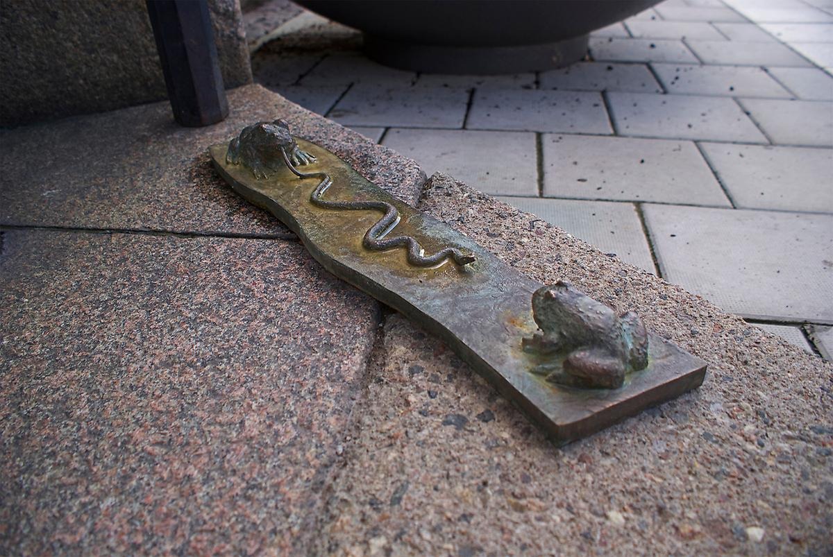 På marken ligger en gråbrun skulptur. Skulpturen föreställer två paddor som sitter mitt emot varandra, paddan till vänster sträcker ut sin tunga som förvandlats till en lång orm som slingrar sig mot den andra paddan. 