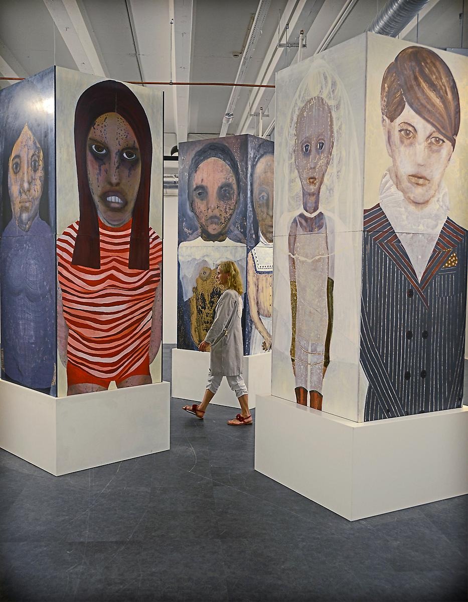 En kvinna går emellan tre stora pelare som är kvadratformade och varje sida består av en oljemålning som föreställer en person. Alla porträtten består av olika personer med olika åldrar, etniciteter och former. 