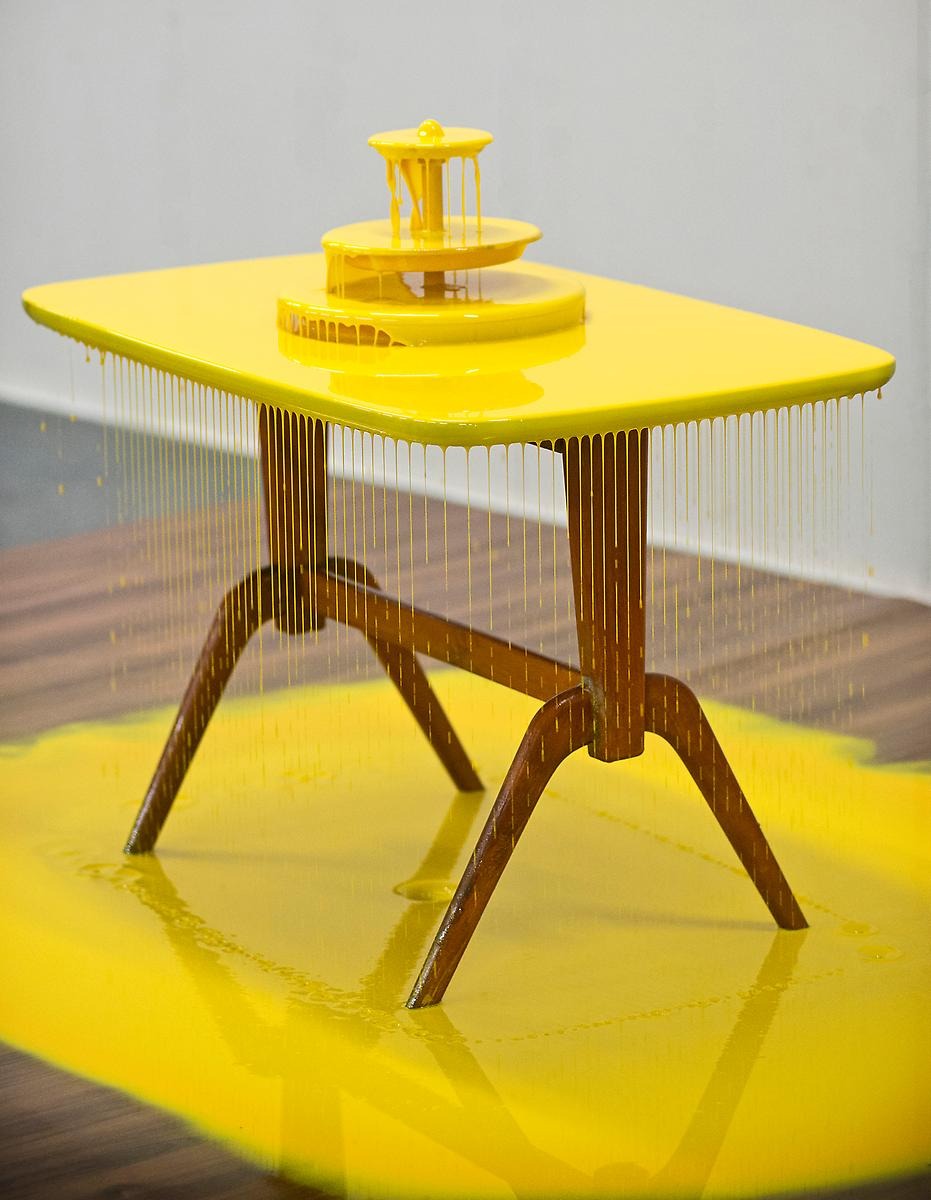 På ett träbord står ett trevåningsfat alla våningar och även bordet är täckt med gul färg som rinner ner från bordets kanter. Även hela golvet under bordet är fyllt med den gula färgen men bordets ben är helt fria.