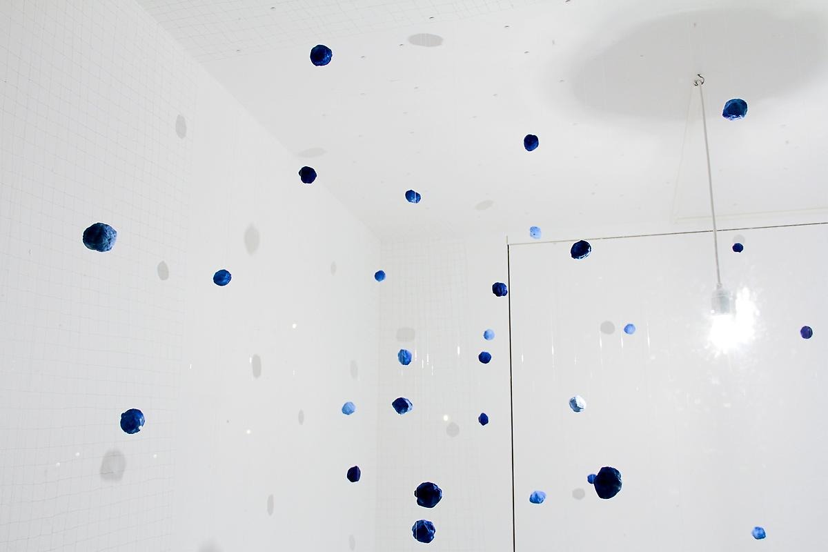 I ett vitt kaklat rum hänger många små blå bollar i olika höjder. Det liknar ett meteoritregn.