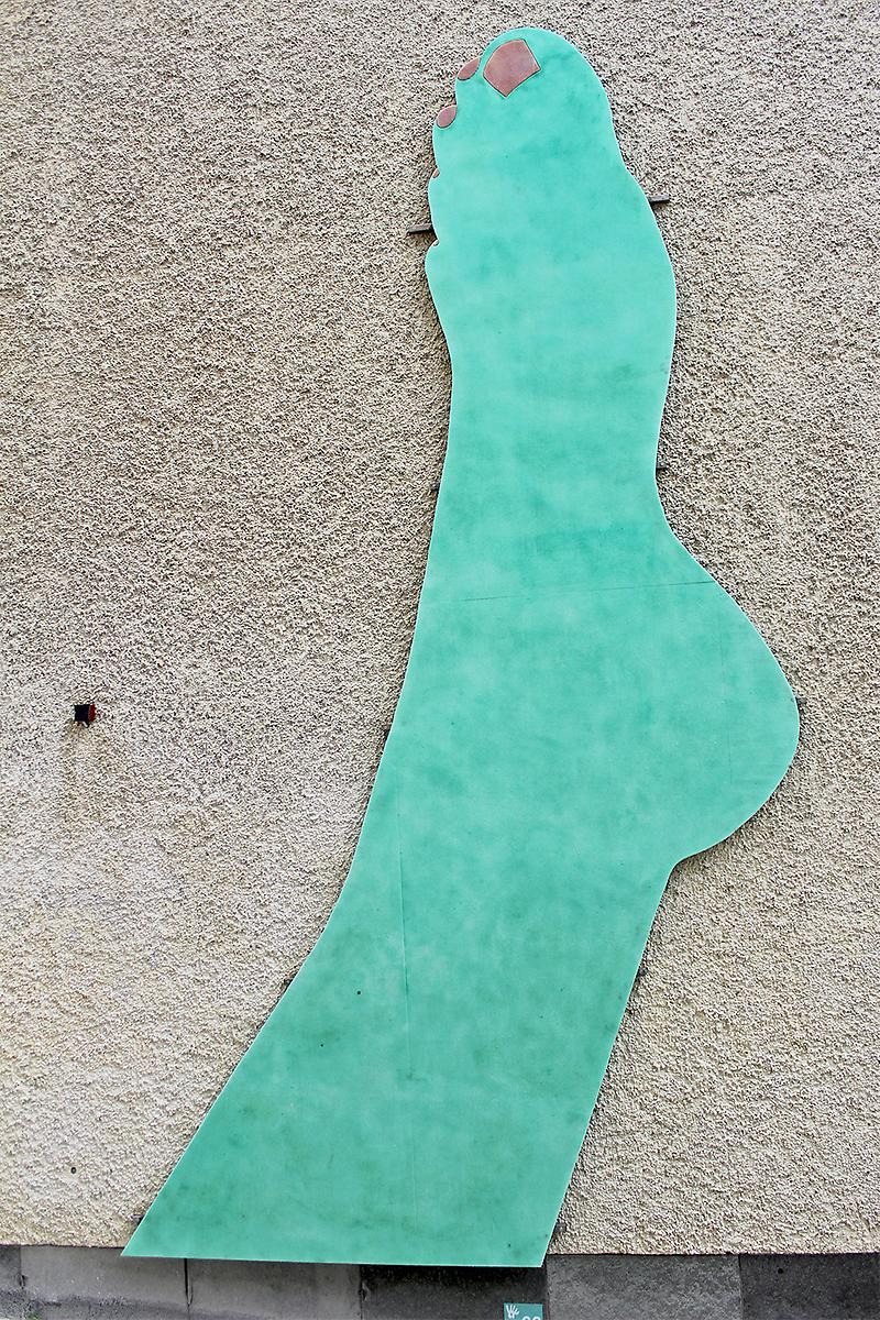 På en husfasad hänger en grön siluett av en fot med bruna tånaglar. 