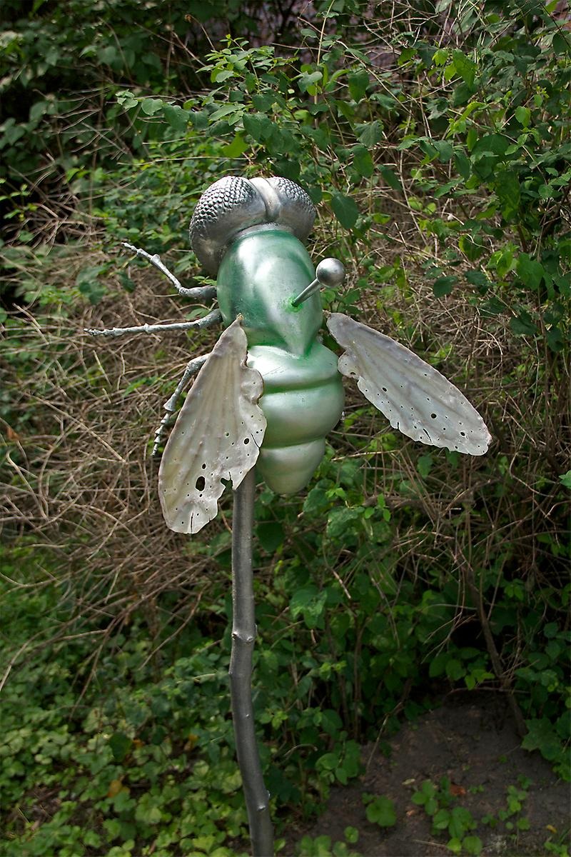I en rabatt sitter en silverfärgad pinne med små knoppar på. Högst upp på pinnen sitter en skimrig fluga med grönfärgad kropp och silverfärgade vingar som har små hål i sig. På mitten av flugans kropp sticker en silvrig nål upp med en knopp.  